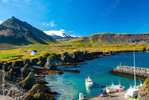 Arnarstapi village in Iceland with Snæfellsjökull volcano in the background, white church on green cliffs above a rocky coastline under a clear blue sky.