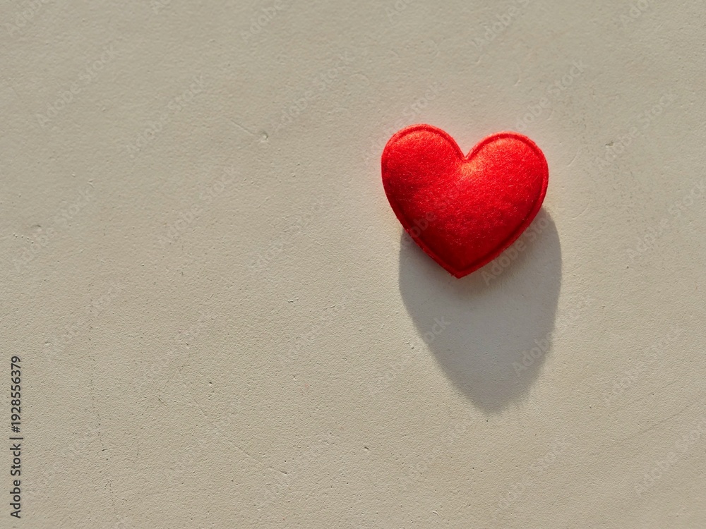 Fototapeta premium Red felt heart on a neutral stone background with sunlight and long shadow. Minimalist romantic concept with a handmade fuzzy wool heart. Top view of a red fiber heart for Valentine's Day, wedding,