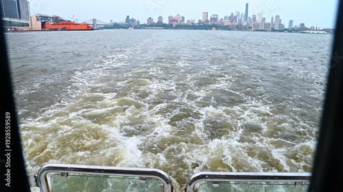 Boat wake on water with Staten Island Ferry background. View from moving vessel showing white water trail with orange ferry and city skyline in distance.