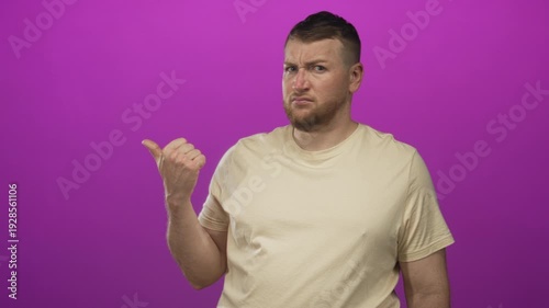 Man pointing thumb to the side in purple studio wearing beige tshirt with scowling face and scrunched nose; skepticism.