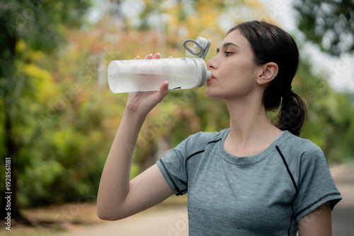 Valokuva Young woman runner holding water bottle in park after jogging, smiling and staying hydrated during outdoor workout, healthy lifestyle and fitness concept