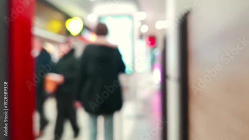 Blurred bokeh footage showing a man silhouette ordering food via bright screen self service terminal inside a shopping mall, with other visitors walking in the soft unfocused background.