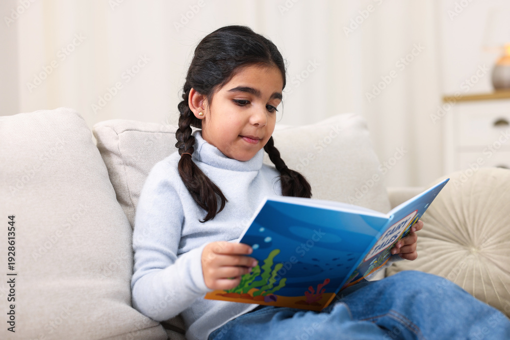 Naklejka premium Cute little girl reading book on couch at home