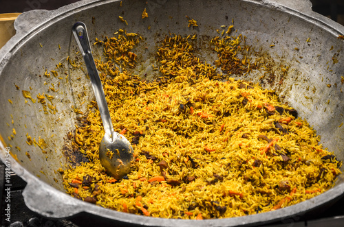 Large metal kazan is filled with pilaf, a seasoned rice dish with pieces of meat and carrots. A silver serving spoon rests inside the pot