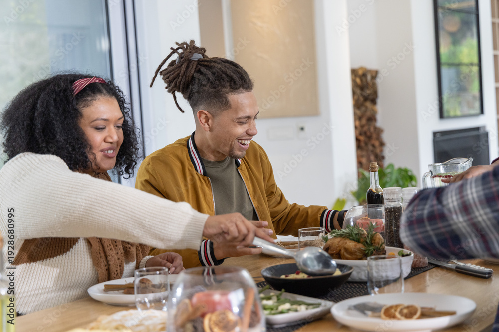 Naklejka premium African American friends smiling and reaching for roasted poultry at wooden dining table in home