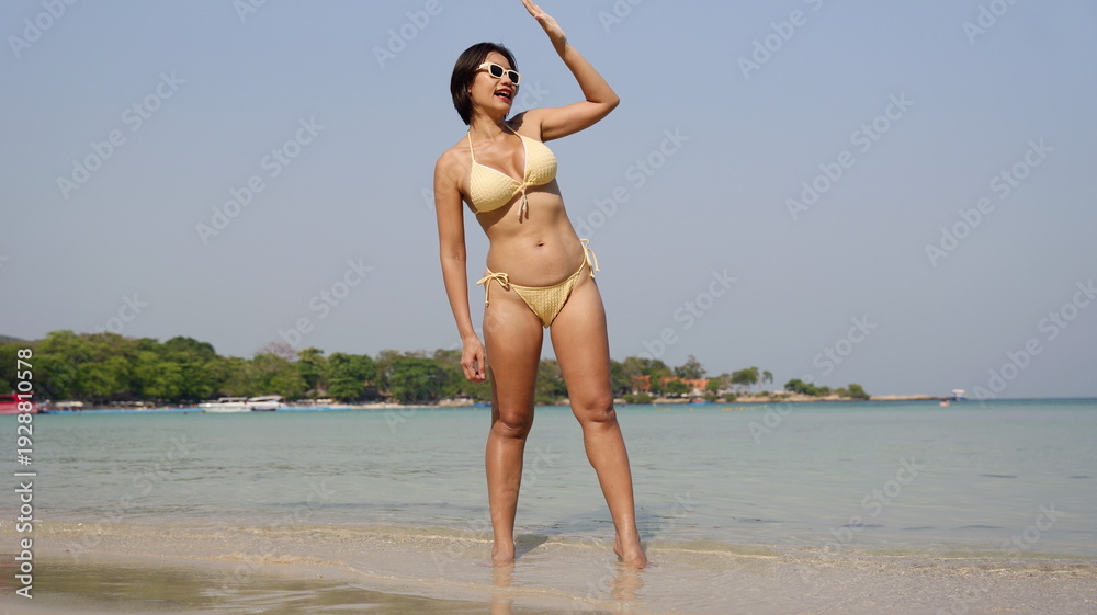 Fototapeta premium A woman in a yellow bikini stands on the shoreline, shielding her eyes from the sun while looking away