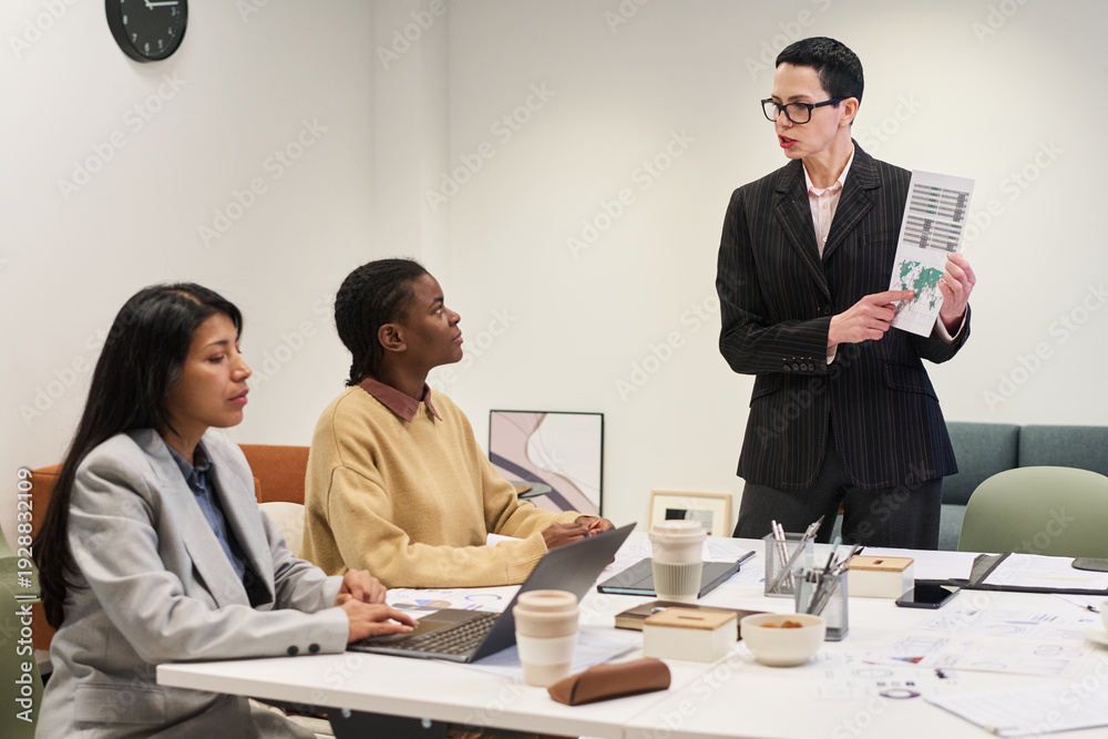 © pressmaster - Middle aged Caucasian woman standing and presenting document to young Black woman and young Hispanic woman sitting at conference table, discussing business strategy during meeting