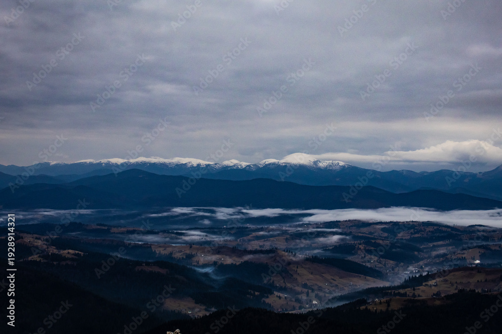 Obraz premium An open panorama shows blue ridges under a dense sky while patches of fog slide across a sprawling valley. Far ranges carry snow and the land below holds scattered villages