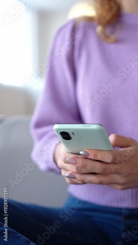 Close-up of lovely blonde teenage girl sitting cross-legged on sofa, chatting online using mobile phone while enjoying leisure and free time in domestic room.