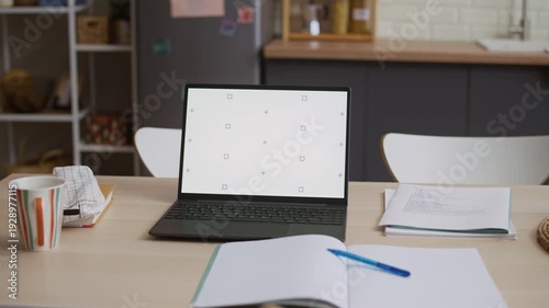 Medium shot of dining light wooden table with opened textbook, papers, stationery and working laptop on top in kitchen area at home