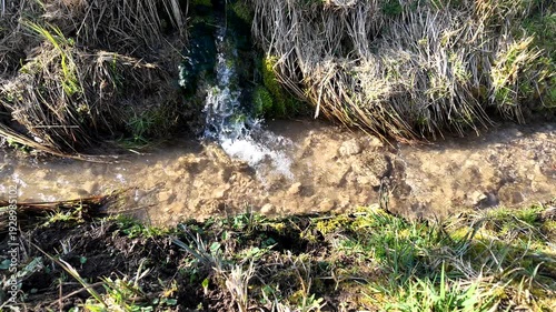 Crystal clear water pours from a small opening into a shallow, stony creek bed surrounded by patches of green grass.