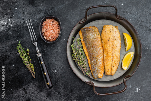 Fotografie Fried sea bass fillets served on ceramic plate with fork and herbs, overhead restaurant style seafood dish ready to eat with dramatic dark backdrop