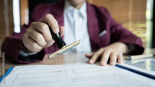 A person is signing a document with a stamp. The stamp is black and has a gold edge. The person is wearing a red suit and white shirt. The document is on a table