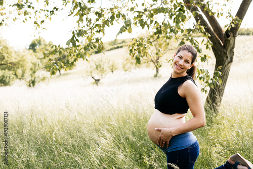 Young pregnant woman holding baby belly, standing on a meadow