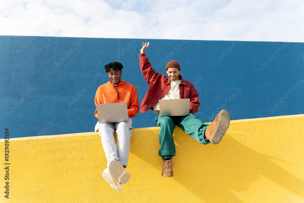 © VISTA by Westend61 - Friends sitting on colorful wall in Barcelona using laptops celebrating success