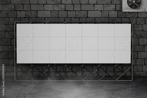 A large, blank, rectangular LED screen is mounted on a metal frame against a dark brick wall, with a small fan visible in the corner