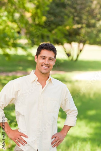 Man standing on grassy lawn in sunlit park, smiling, posing hands on hips wearing light shirt