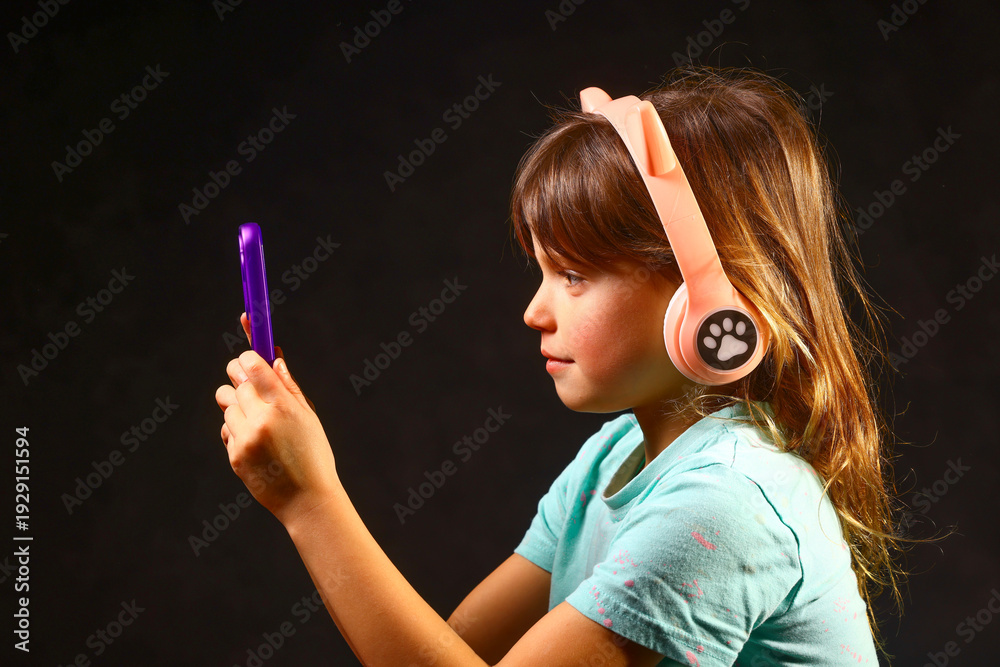 Obraz premium Young child focused on a smartphone while wearing playful wireless headphones, showing how modern technology blends naturally into everyday moments of curiosity and early digital exploration