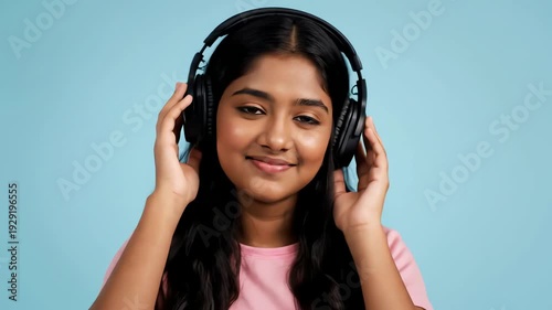 Smiling Young Woman With Black Wavy Hair Wearing Black Headphones and a Pink Shirt Against a Light Blue Background
