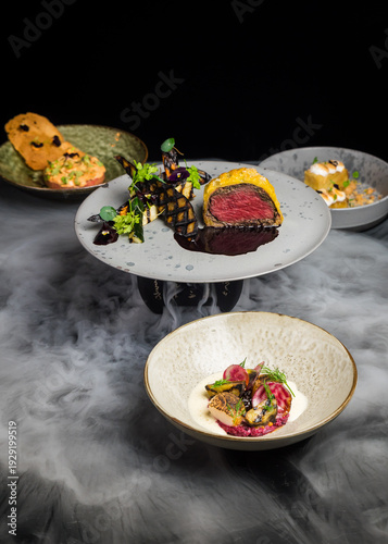 A mix of gourmet fusion fine dining dishes, including beef wellington, steak tartare, scallops and a mango cake dessert with dry ice creating a mist or smoke like effect