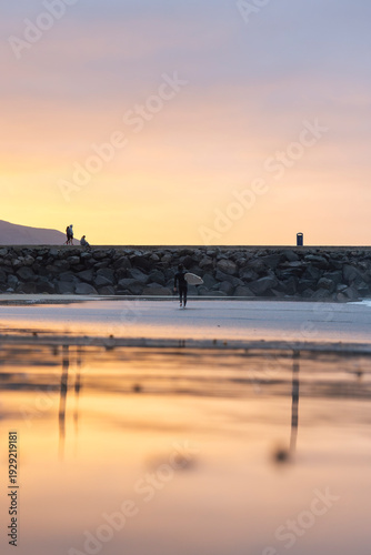 Silhouette of a person with a surfboard along a coastal breakwater at sunset, with gentle waves washing ashore under a warm golden sky