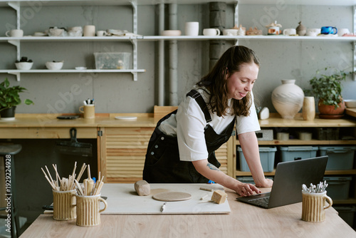 Fotografia Young adult Caucasian woman standing at pottery studio table using laptop, surro