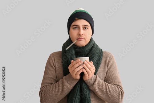 Ill young man with cup of tea and thermometer measuring temperature on light ...