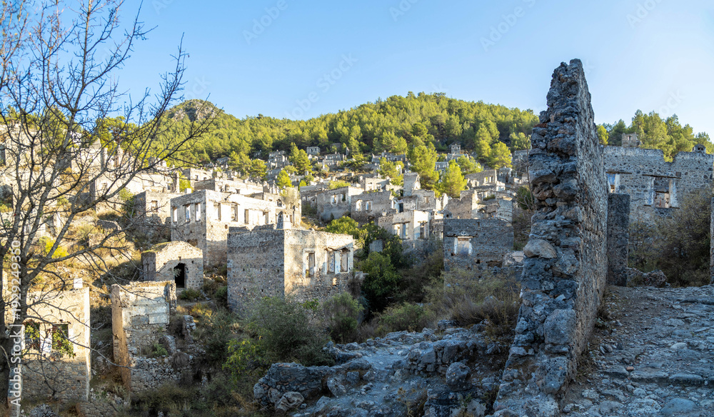 Fototapeta premium Panoramic view of Kayakoy ghost town with abandoned stone houses. Ancient ruins of a greek village on a green hill near Fethiye, Mugla province, Turkey. Sunny autumn day in October.