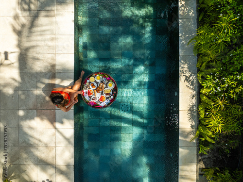 Aerial view of a woman lounging with a floating breakfast tray in the pool, framed by the geometric tiles and lush greenery, Prince Maurice, Flacq District, Mauritius.