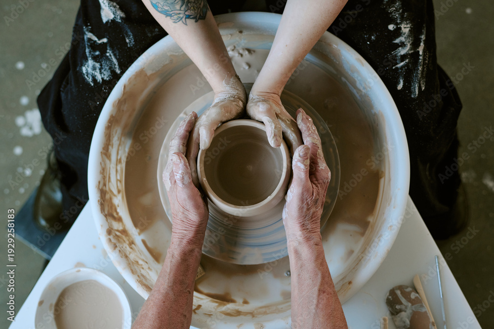 © AnnaStills - Unrecognizable senior mother guiding young adult daughter shaping clay on pottery wheel, both hands covered in wet clay, close up of collaborative creative process