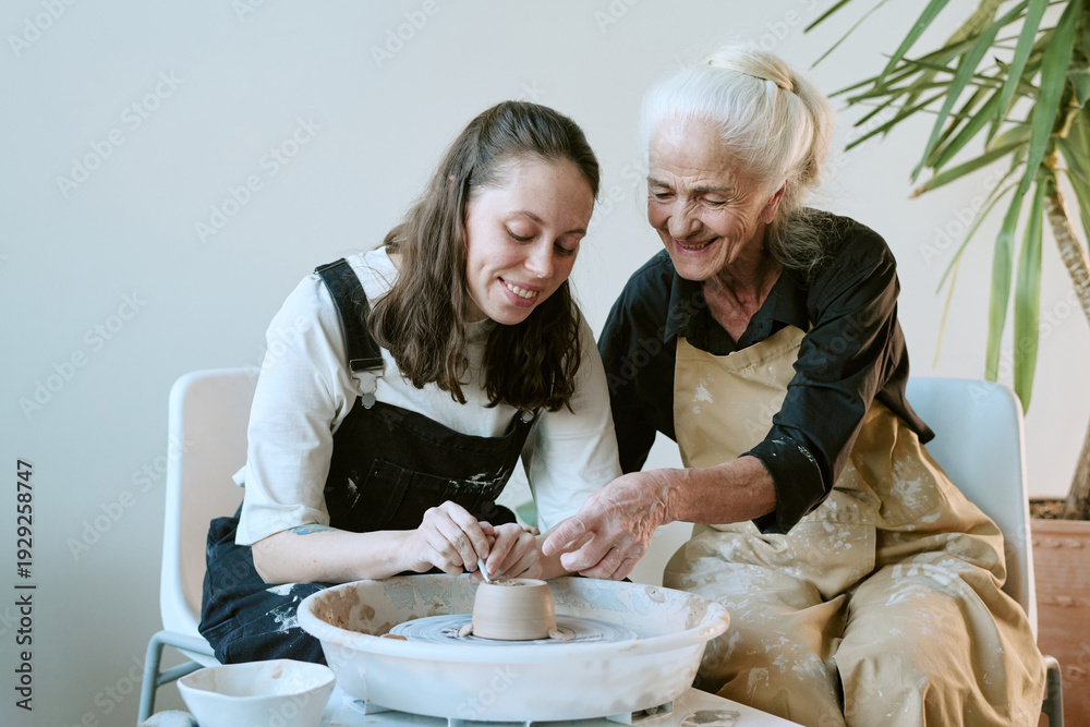© AnnaStills - Caucasian young adult woman and Caucasian senior woman shaping clay together on pottery wheel, both smiling and focusing on hands while creating ceramic piece in studio setting © AnnaStills - Caucasian young adult woman and Caucasian senior woman shaping clay together on pottery wheel, both smiling and focusing on hands while creating ceramic piece in studio setting