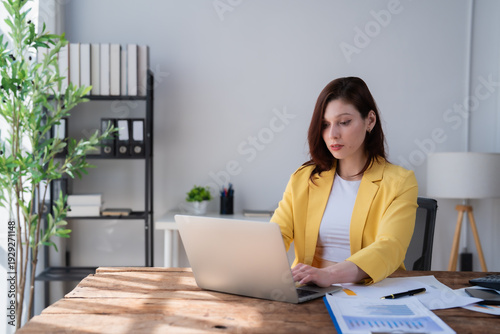 Foto Woman in a yellow blazer focusing on her laptop, analyzing financial documents a