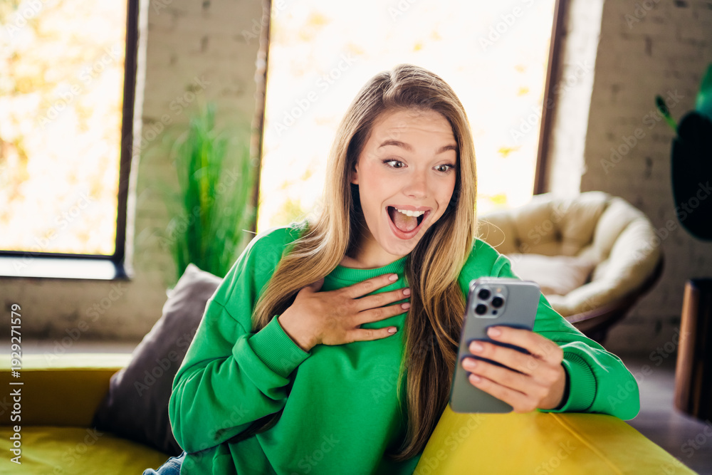 Fototapeta premium Joyful young woman in green sweatshirt reacting to exciting news with her smartphone in a cozy interior loft