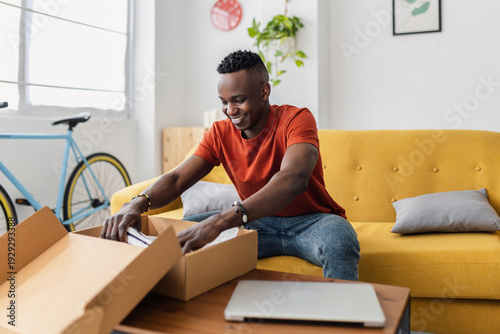 Obraz na płótnie Happy African man opening delivery box with new clothes purchased online at home
