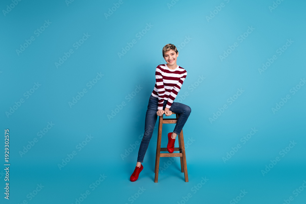 Fototapeta premium Young woman sits on a wooden stool against a bright blue background wearing a striped sweater jeans and red shoes posing for a stylish fashion lifestyle shoot