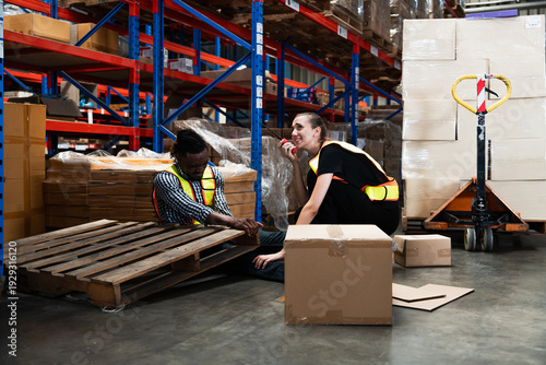 African American warehouse worker kneeling to assist injured Caucasian female colleague holding ankle after minor accident on concrete floor, surrounded by boxes.
