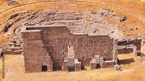 Cinematic 4K drone shot of the ancient Roman theater of Acinipo archaeological site, showing the standing stone wall and seating area in the mountains near Ronda, Malaga province.