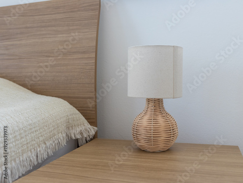 Minimalist bedside lamp with rattan base on a wooden nightstand. Modern boho bedroom interior with cozy lighting and natural textures.