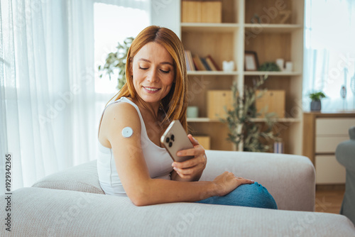 Bild auf Leinwand Woman Checking Glucose Monitor With Smartphone App While Relaxing On Couch At Ho