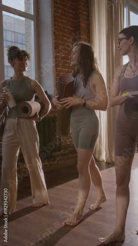 Full vertical tracking shot of three sporty young Caucasian female friends with yoga mats walking and chatting in corridor of loft yoga studio to group class
