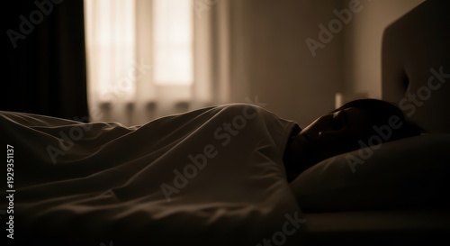 A women lies in bed with their head on a pillow, covered by a blanket, in a dark room with a window in the background.