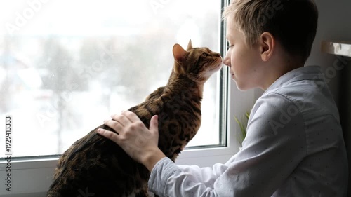 Boy Kissing and Petting Bengal Cat on Windowsill, Snow Falling Outside