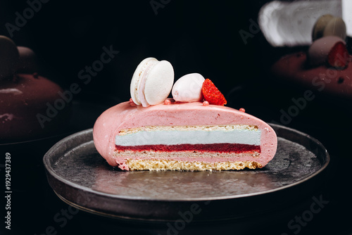 Slice of pink strawberry mousse cake with macarons and berry garnish on metal plate