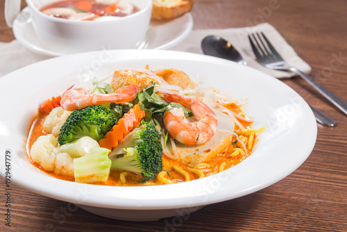 A white plate of spicy Thai noodle soup topped with shrimp, broccoli, carrots, and fresh herbs, served with soup and bread on the side.