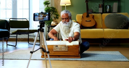 Indian mid age bearded man recording harmonium playing session on smartphone for online class at home, stylish mature music tutor creating classical lesson video with tripod setup in modern studio