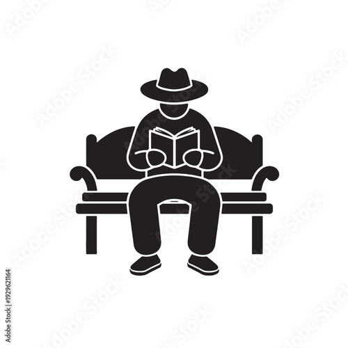 A smiling senior businessman is sitting on a chair at an office desk reading a book near his laptop while a professional woman worker completes her job at a table