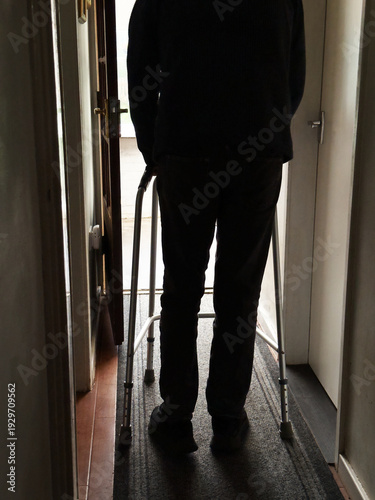 A slim adult in silhouette holding a lightweight walking frame as he moves along a dimly lit indoor passage or corridor, towards a lighter area.