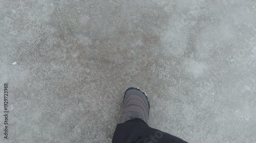 A top-down view of a person wearing grey winter boots walking on a textured icy ground. Close up.
