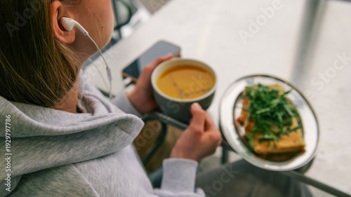 Young woman wearing headphones enjoys a meal of soup and sandwich while listening to music