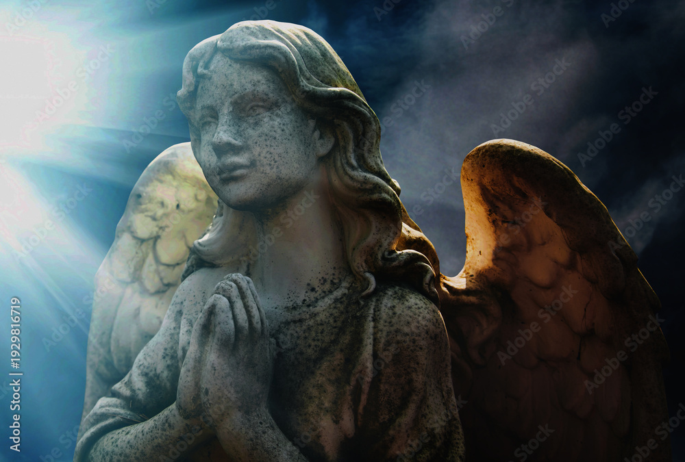 Obraz premium Angel statue in prayer illuminated by heavenly light rays, symbol of hope, protection, spirituality, and divine presence.